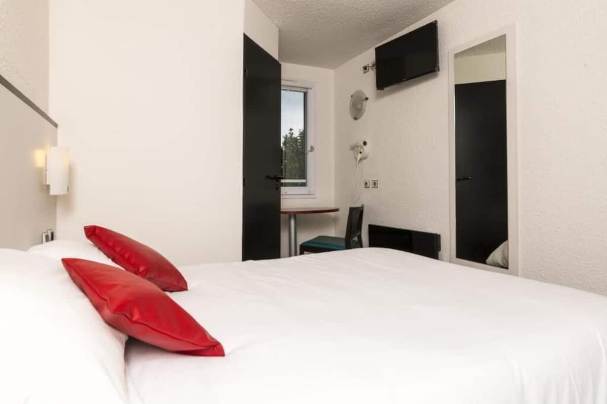 A hotel room with a bed, two red pillows, a desk with a chair, a TV mounted on the wall, and a window.