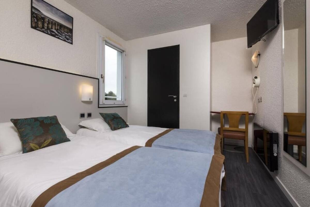 A hotel room with two beds, a desk, a chair, a TV, and a window.