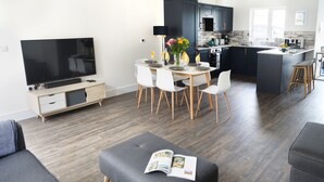 A modern living room with a flat-screen TV, a dining area with a table and chairs, a grey ottoman with a magazine, and a kitchen with dark cabinets and a window.