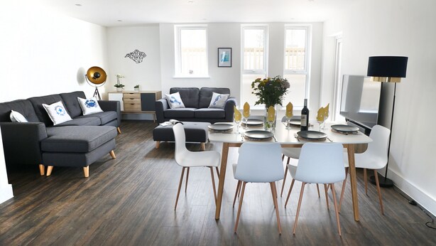 A modern living room with a grey sectional sofa, a dining table with white chairs, and a flat-screen TV.