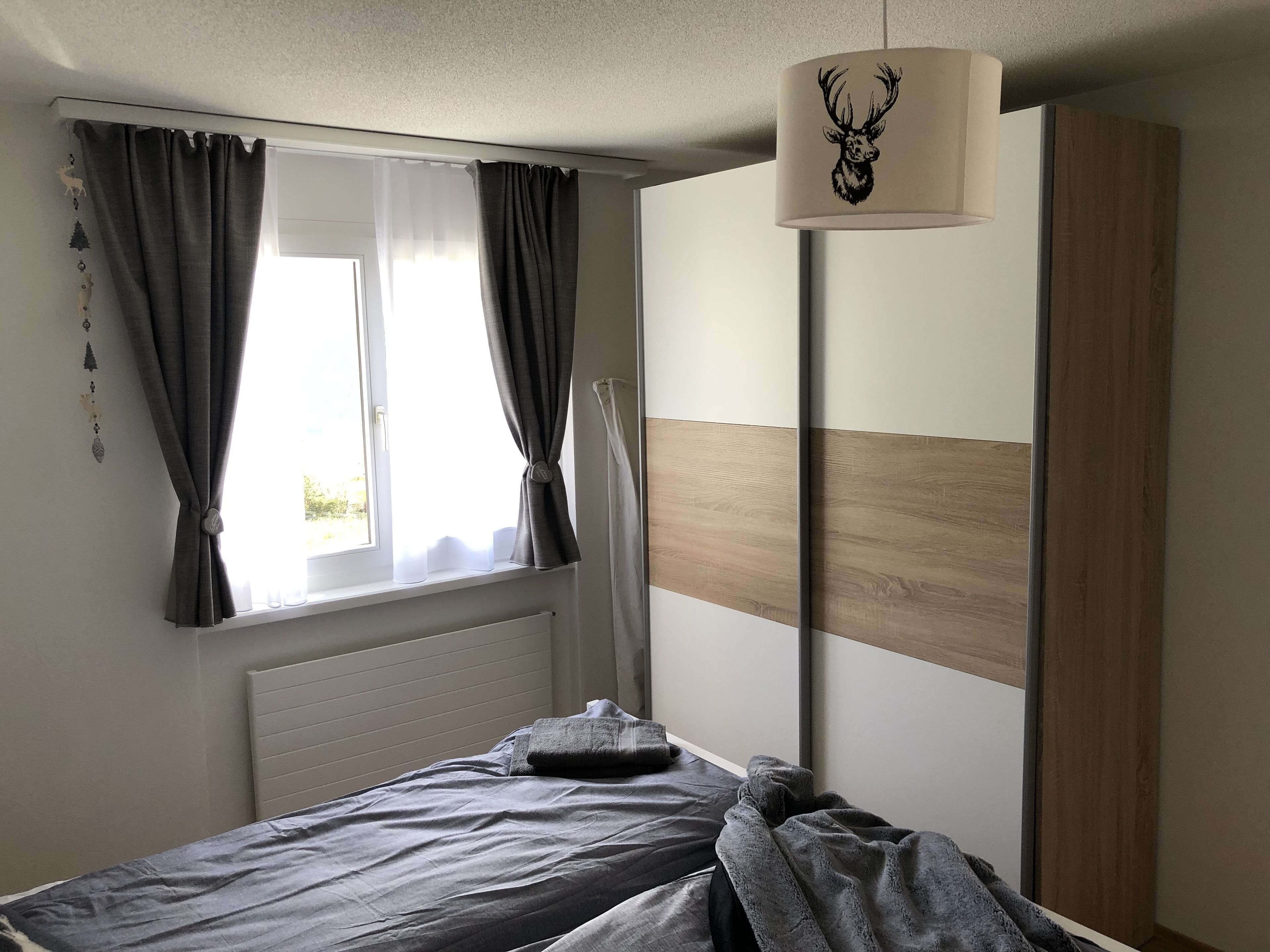 A bedroom with a bed, a window with curtains, a radiator, and a wardrobe. - Renovated 3 Rooms Apartment in Ruschein Near Laax (Ilanz)
