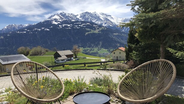 A patio with two wicker chairs and a small table, overlooking a mountain landscape with houses and a clear sky. - Renovated 3 Rooms Apartment in Ruschein Near Laax (Ilanz)