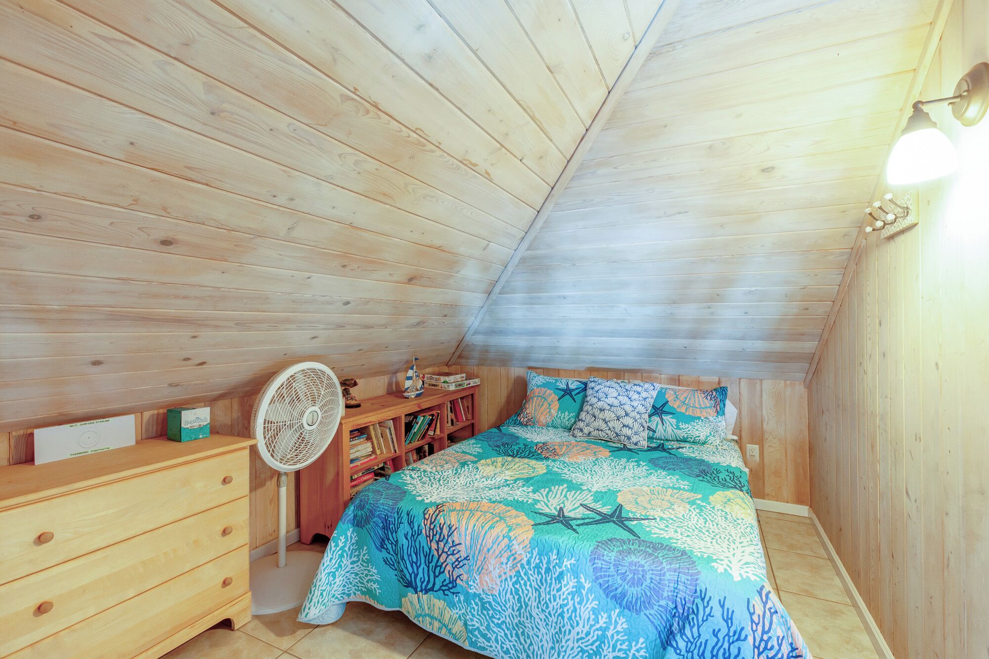 Ein gemütliches Schlafzimmer im Dachgeschoss mit Holzbalkendecke, einem Bett mit Bettdecke in blau-weißem Korallmuster, einer Holzkommode, einem Ventilator und einem Regal mit Büchern und einem kleinen Schiffmodell.
