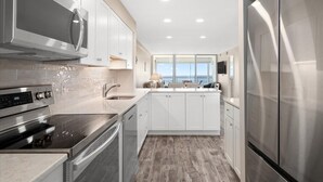A modern kitchen with stainless steel appliances, white cabinetry, and a large window offering a view of the ocean. - English Towers-Beautiful Direct Ocean Views from this Remodeled Condo! (Ocean City)