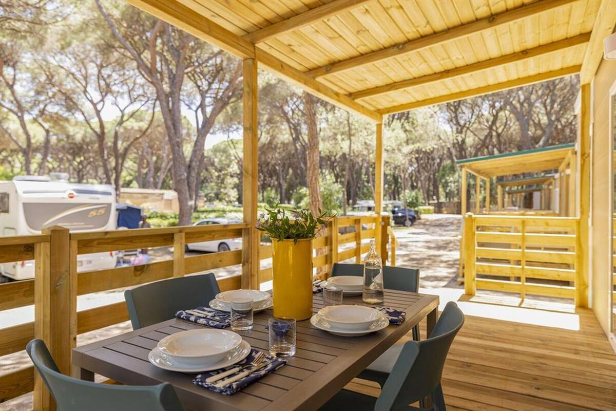 Un área de comedor al aire libre techada con una mesa de madera dispuesta para dos personas, rodeada de sillas y una maceta amarilla con plantas.