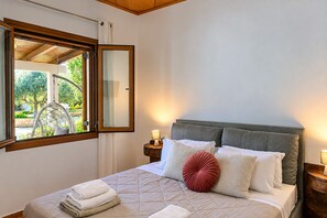 A bedroom with a bed, a nightstand, a lamp, and a window with a view of greenery. - Villa Avra - With Private Pool (Rethymno)