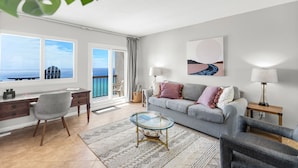 Uma sala de estar moderna com sofá, poltronas, mesa de centro de vidro e escrivaninha. Há uma varanda com vista para o oceano.