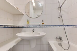 Ein Badezimmer mit einem weißen Waschbecken, einem Spiegel, einer Glasablage mit Pflegeprodukten und einer Dusche.