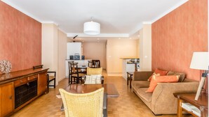 A living room with a sofa, dining table, and chairs, a kitchen area with a refrigerator and microwave, and a hallway leading to a bedroom.