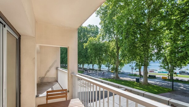A balcony with a wooden table and chairs, overlooking a street with trees and a body of water.