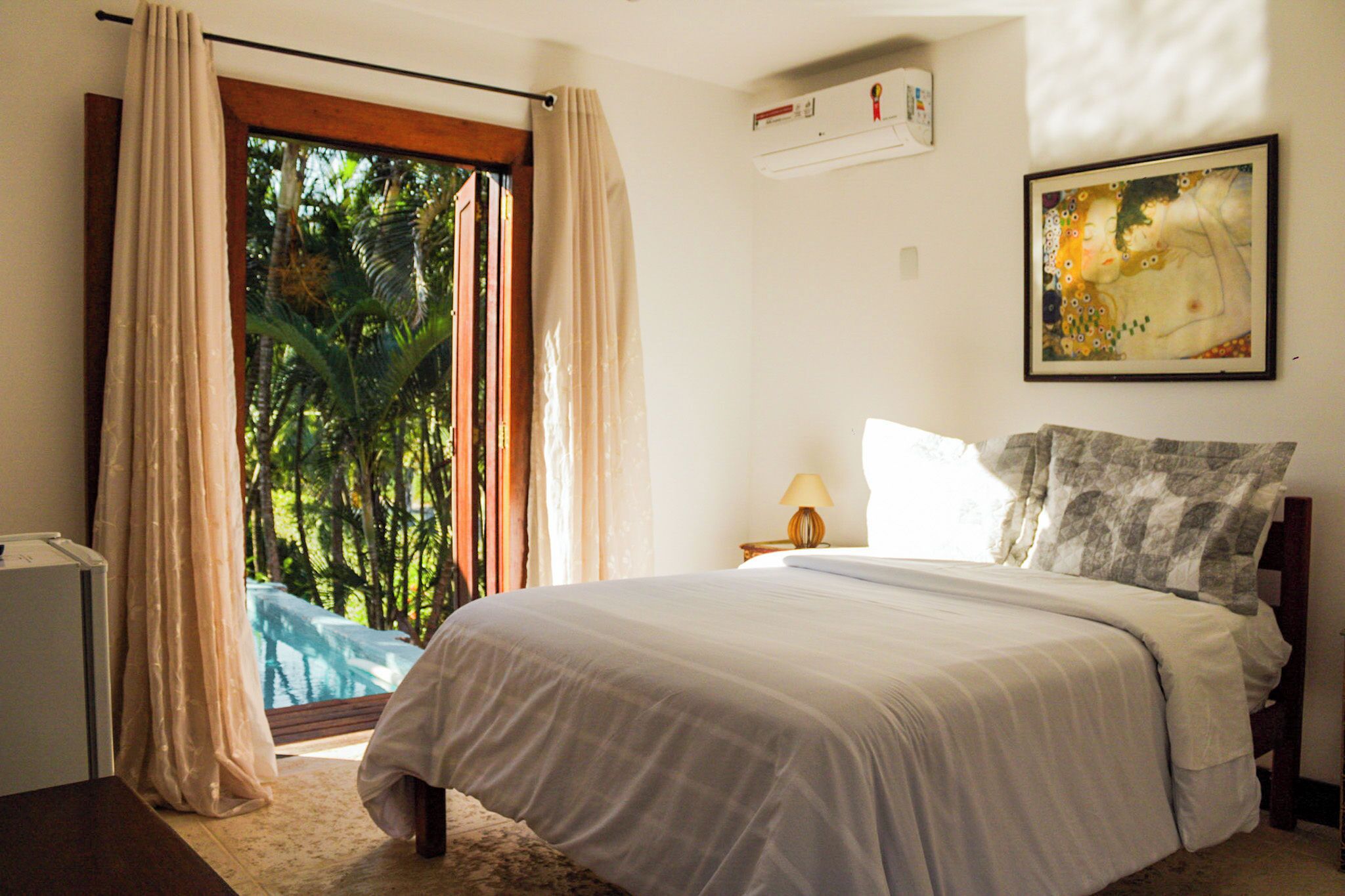 A bedroom with a bed, a nightstand, a lamp, a painting, and a view of greenery through a window.