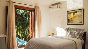 Un dormitorio con cama, mesita de noche, lámpara, un cuadro y vistas al verdor a través de una ventana.