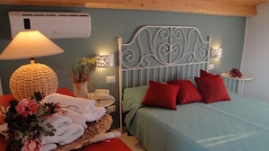 Un dormitorio con cabecera de metal, una cama con almohadas rojas, una mesita de noche con lámpara y toallas, y un aire acondicionado instalado en la pared.