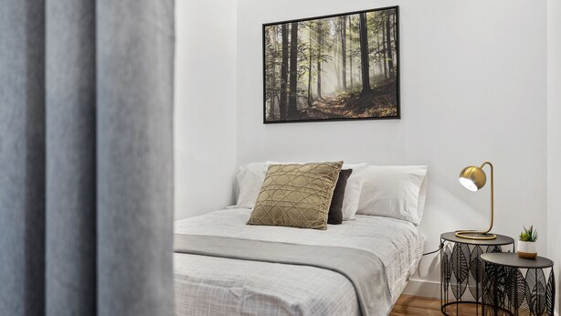 Een slaapkamer met een bed, een nachtkastje met een lamp, een wanddecoratie van een bos en een kleine potplant.