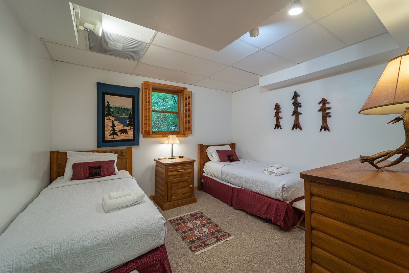 Quarto com duas camas de solteiro, mesa de cabeceira, luminária, janela com persianas e um enfeite na parede.