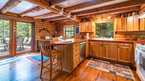 A rustic cabin interior with a wooden dining table, chairs, a kitchen with wooden cabinets, and a large window. - 3BR cottage on the Toccoa River; fish from the yard, stargaze at the firepit! (Suches)