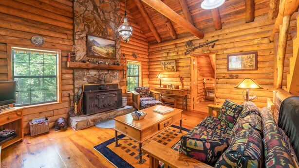 A cozy log cabin interior with a stone fireplace, wooden furniture, and a large window overlooking the forest. - 3BR cottage on the Toccoa River; fish from the yard, stargaze at the firepit! (Suches)