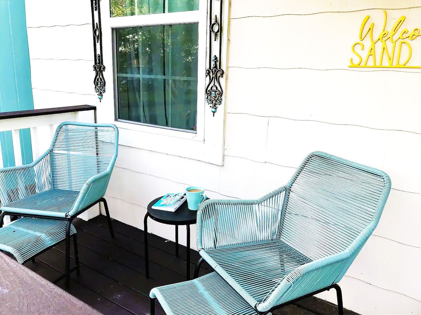 A porch with two turquoise wicker chairs, a small table with a cup, and a 'Welcome Sand' sign on the wall.