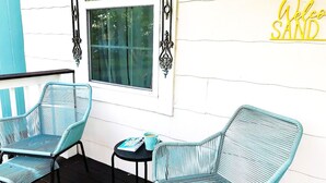 Une véranda avec deux fauteuils en osier turquoise, une petite table avec une tasse, et une pancarte « Bienvenue à Sand » accrochée au mur.