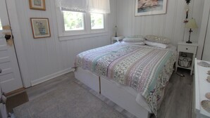 Un dormitorio con cama, mesita de noche, cómoda, ventana con cortinas y cuadros en la pared.