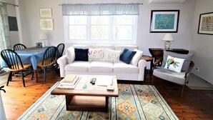 Una sala de estar con un sofá blanco, una mesa de centro de madera, un tapete estampado y un comedor con una mesa y sillas.