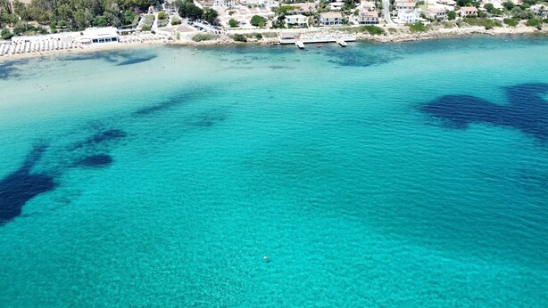 Une zone côtière aux eaux turquoise et limpides, une plage parsemée de parasols et un port de plaisance où sont amarrés des bateaux.