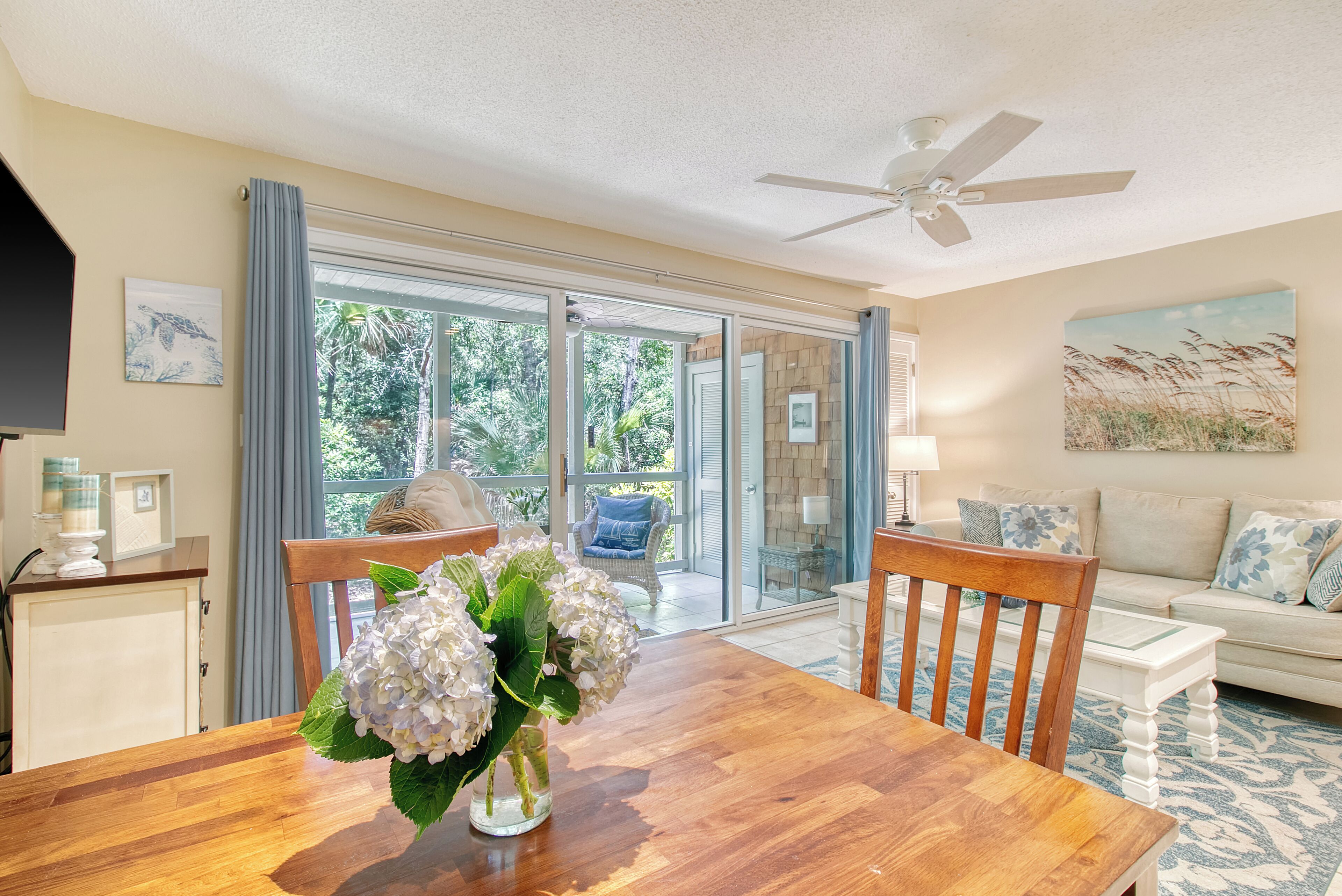 A dining area with a wooden table, chairs, and a vase of flowers. There is a ceiling fan, a sofa, and a painting on the wall.