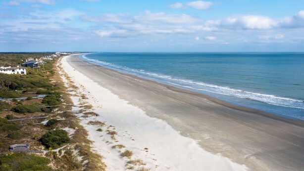 A coastal beach with a long stretch of sand, a few buildings along the shore, and a clear view of the ocean. - Across from beach, new bathroom reno! (Kiawah Island)