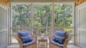 A room with two wicker chairs, a small table, and a view of a forest through large windows. - Across from beach, new bathroom reno! (Kiawah Island)