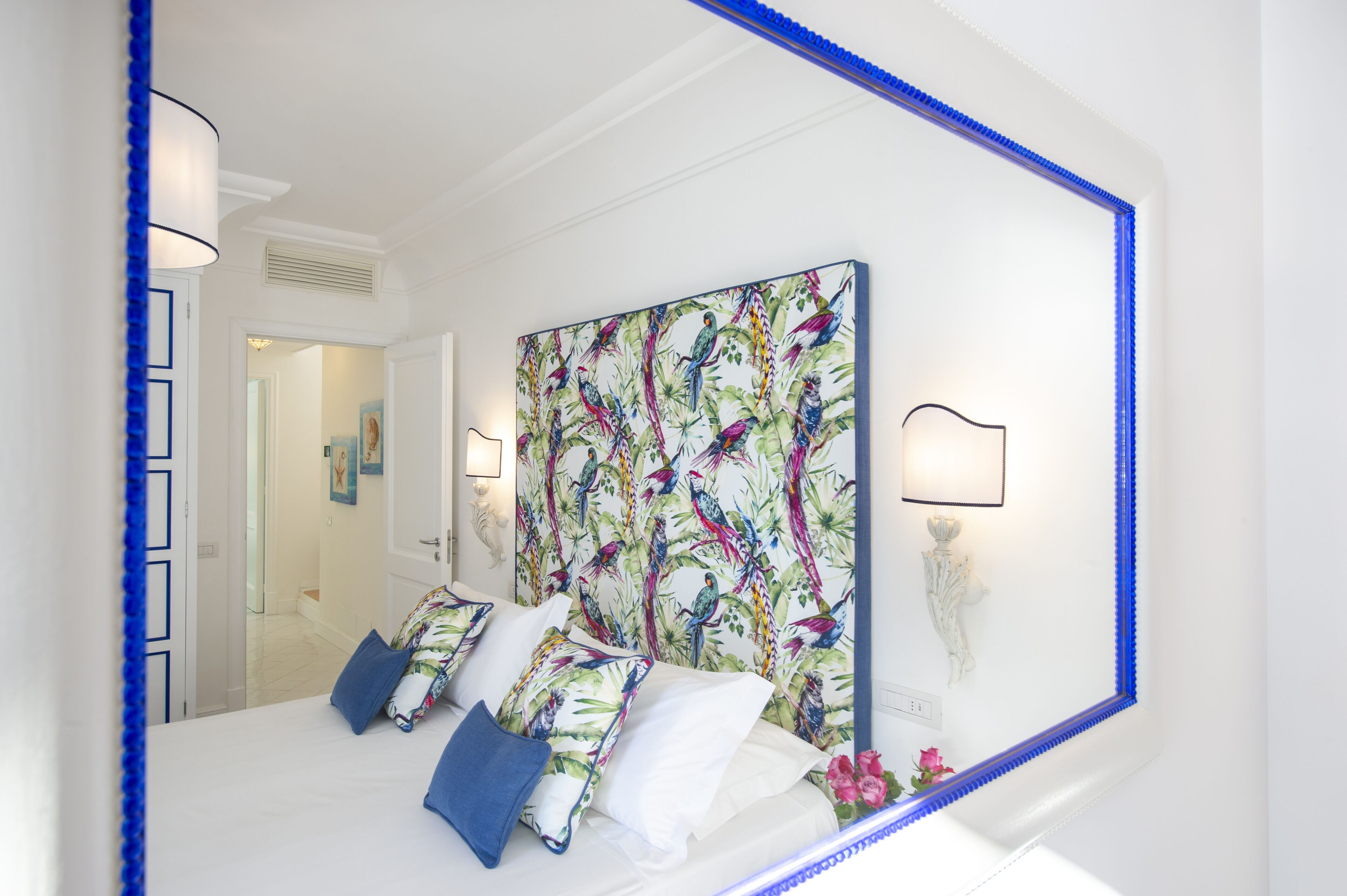 Ein Schlafzimmer mit einem großen Bett, einem farbenfrohen Wandbehang zum Thema Vögel und einem Spiegel mit blauem Rahmen.