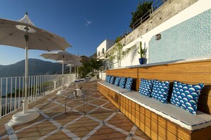 Une terrasse dotée d’un sol carrelé, de parasols blancs, d’un banc en bois avec des coussins à motifs bleus et d’un mur orné de motifs.