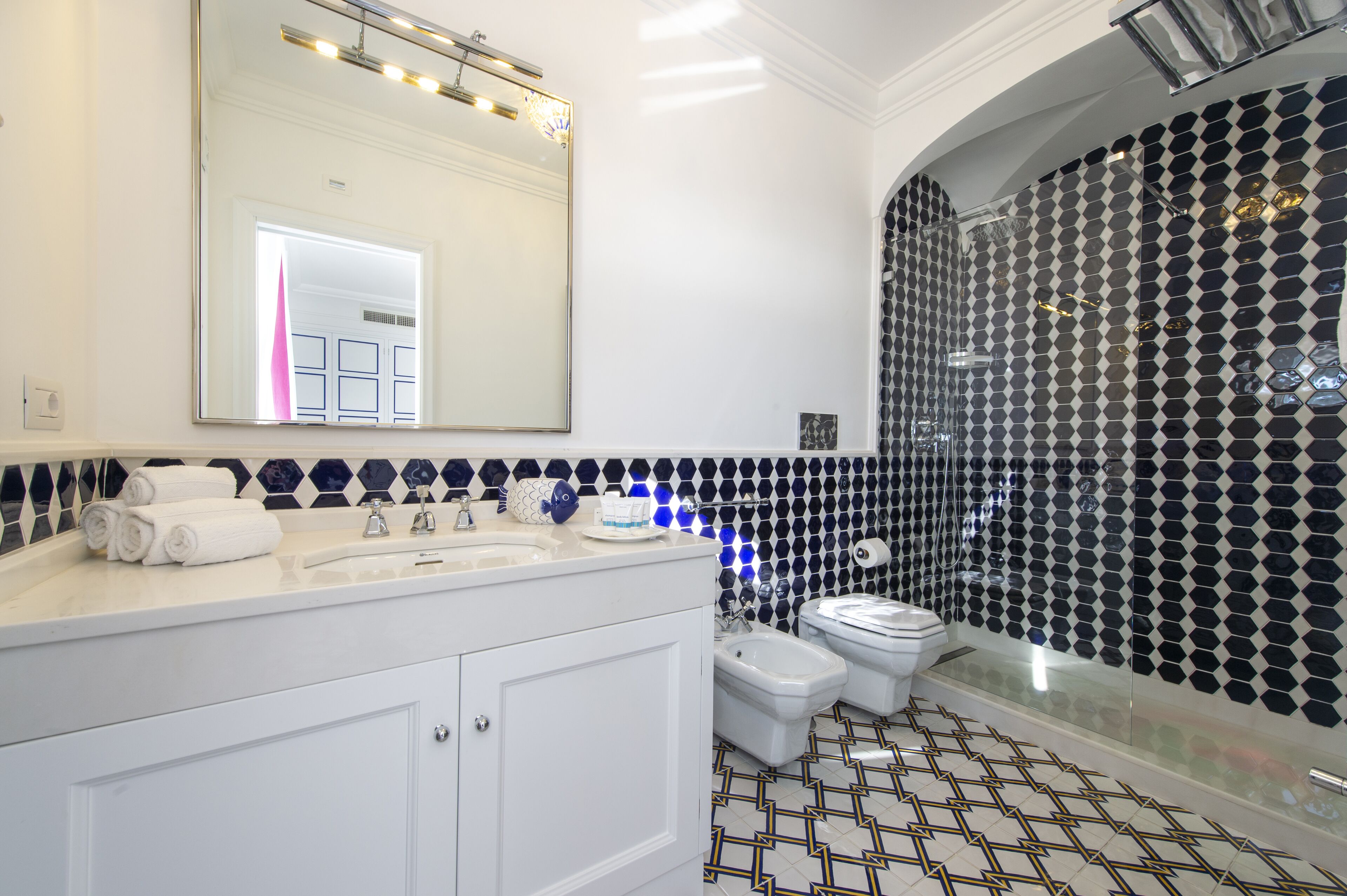 Une salle de bain avec un meuble-lavabo blanc, un grand miroir, une douche avec un mur orné d’un motif noir et blanc, et un sol carrelé.