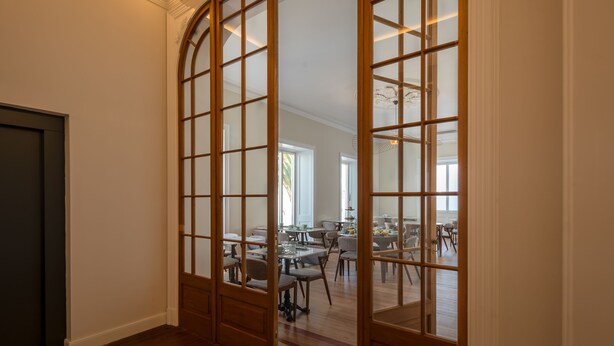 Quarto com divisória de madeira e painéis de vidro, área de refeições com mesas e cadeiras, e vista para uma cozinha ao fundo.