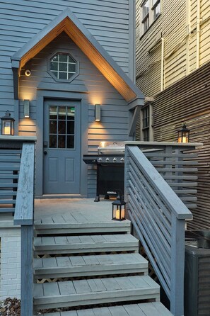 A house with a grey exterior, a wooden staircase leading to a front door, and a barbecue grill outside.