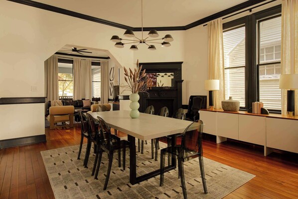 A dining area with a long table, chairs, and a chandelier. There is a fireplace and a sofa in the background.