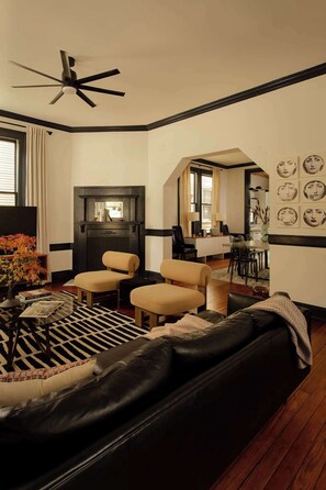 A living room with a black leather sofa, two beige armchairs, a coffee table with a vase and books, a fireplace, and a dining area in the background.