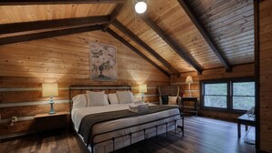 Quarto acolhedor em cabana de madeira, com uma cama, duas mesas de cabeceira, uma cadeira e uma janela grande.