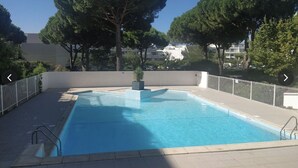 Uma piscina retangular com um deck ladrilhado, cercada por uma vedação branca, e árvores ao fundo.