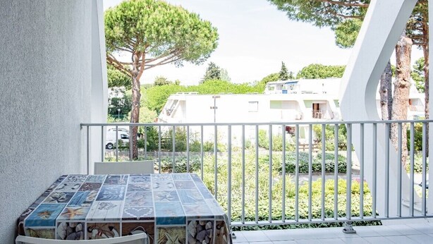 Uma varanda com mesa e cadeiras azulejadas, grade branca e vista para edifícios e árvores.