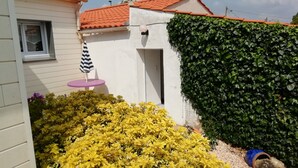 A white building with a red-tiled roof, a small window, and a door. There is a purple table with a striped umbrella, yellow flowering bushes, and a green ivy-covered wall.