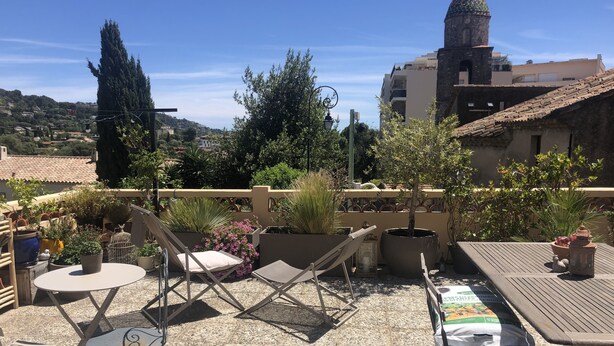 Un patio al aire libre con mobiliario, plantas en macetas y vistas a un pueblo con una torre destacada.