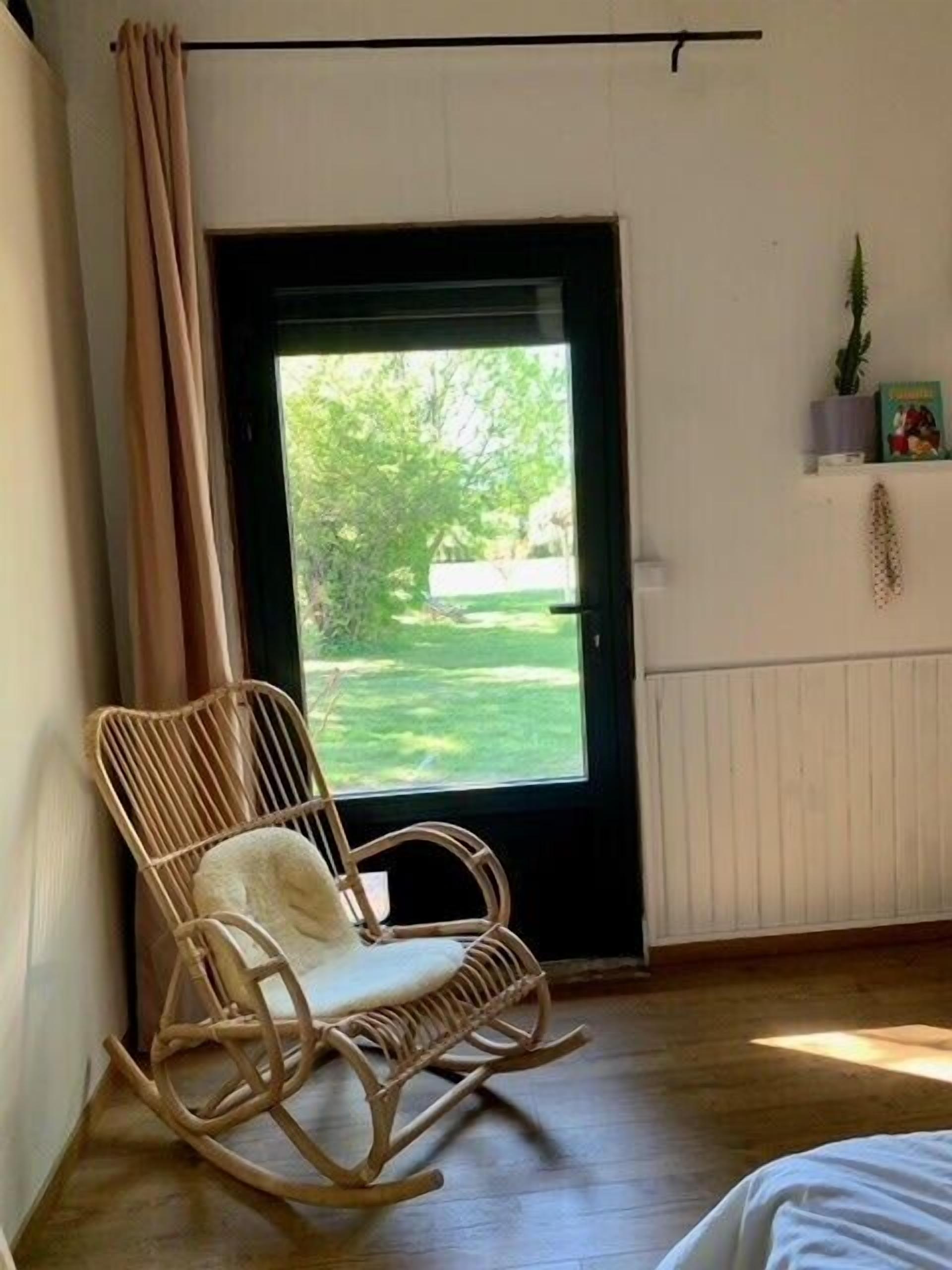 A room with a rocking chair, a bed, a window with a view of greenery, and a shelf with a plant and a book.
