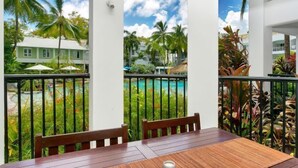 Ein Holztisch mit zwei Stühlen auf einem Balkon mit Blick auf einen Swimmingpool und Palmen.