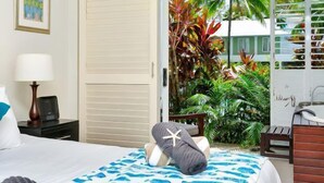 A hotel room with a bed, a desk, a lamp, a chair, and a view of tropical plants through an open door. - Ground floor Spa Suite in the popular Beach Club Resort in Palm Cove (Palm Cove)