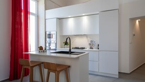 Uma cozinha moderna com armários brancos, uma ilha central com banquetas, forno embutido e uma cortina vermelha.