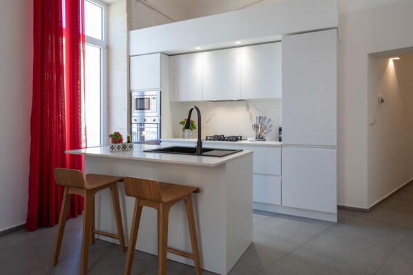 Een moderne keuken met witte kasten, een centraal eiland met barstoelen, een inbouwoven en een rood gordijn.