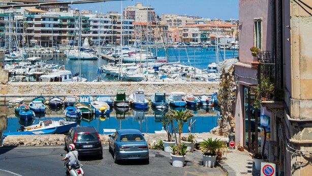 Uma marina com barcos atracados, uma rua com carros e um motociclista, e edifícios ao fundo.