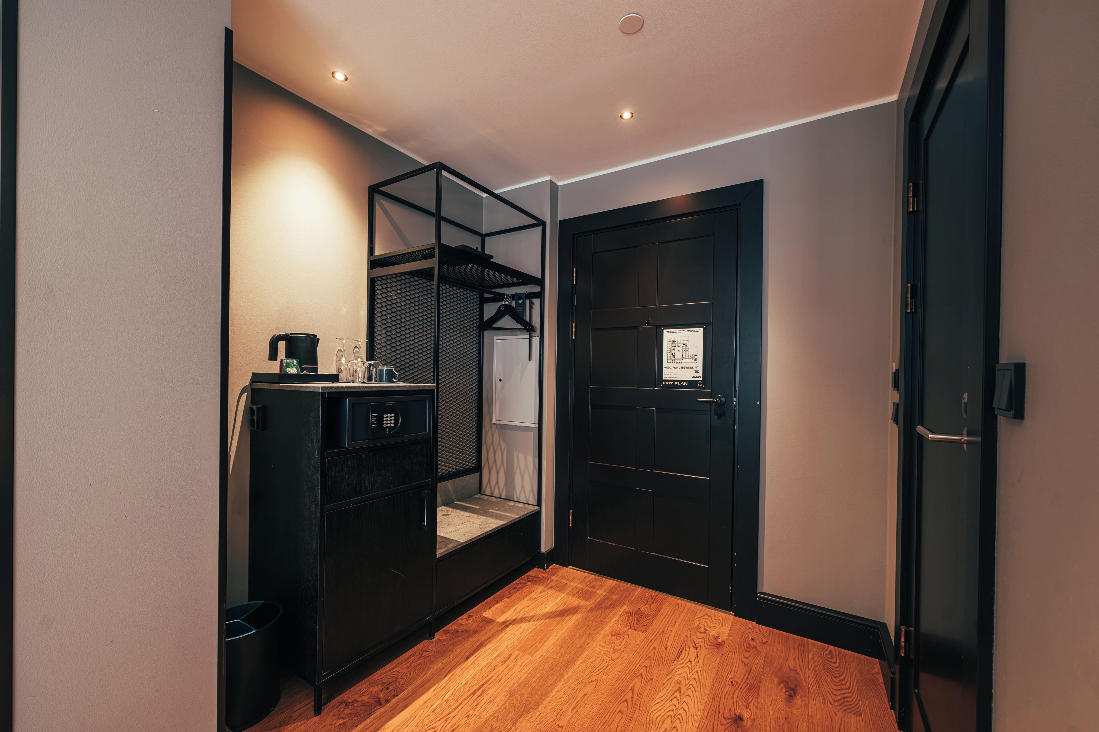 A compact room with a wooden floor, a black metal cabinet with a hanging rack, a door with a security lock, and a small trash bin.