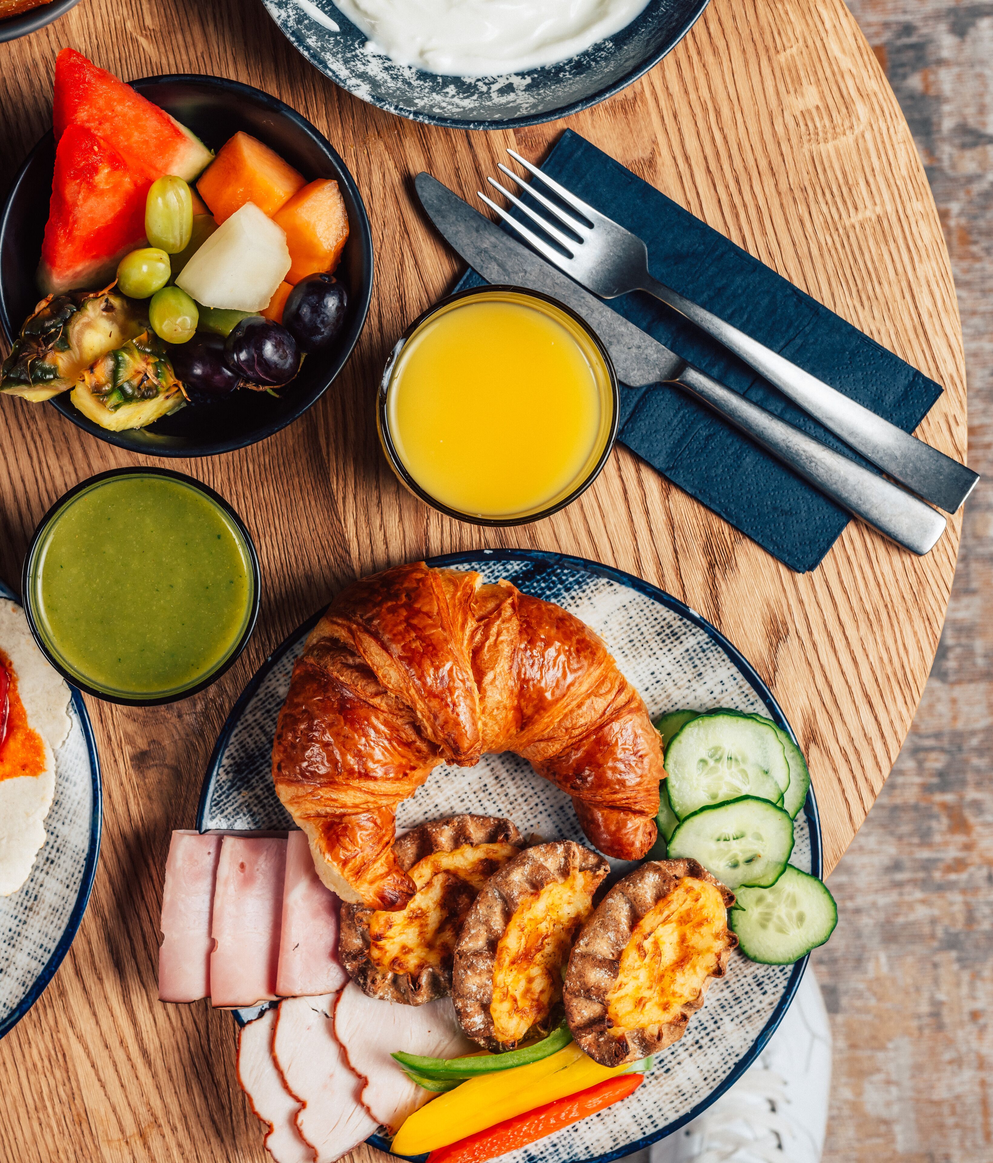 A breakfast setting with croissants, ham, grilled sausages, sliced cucumbers, and a fruit bowl.