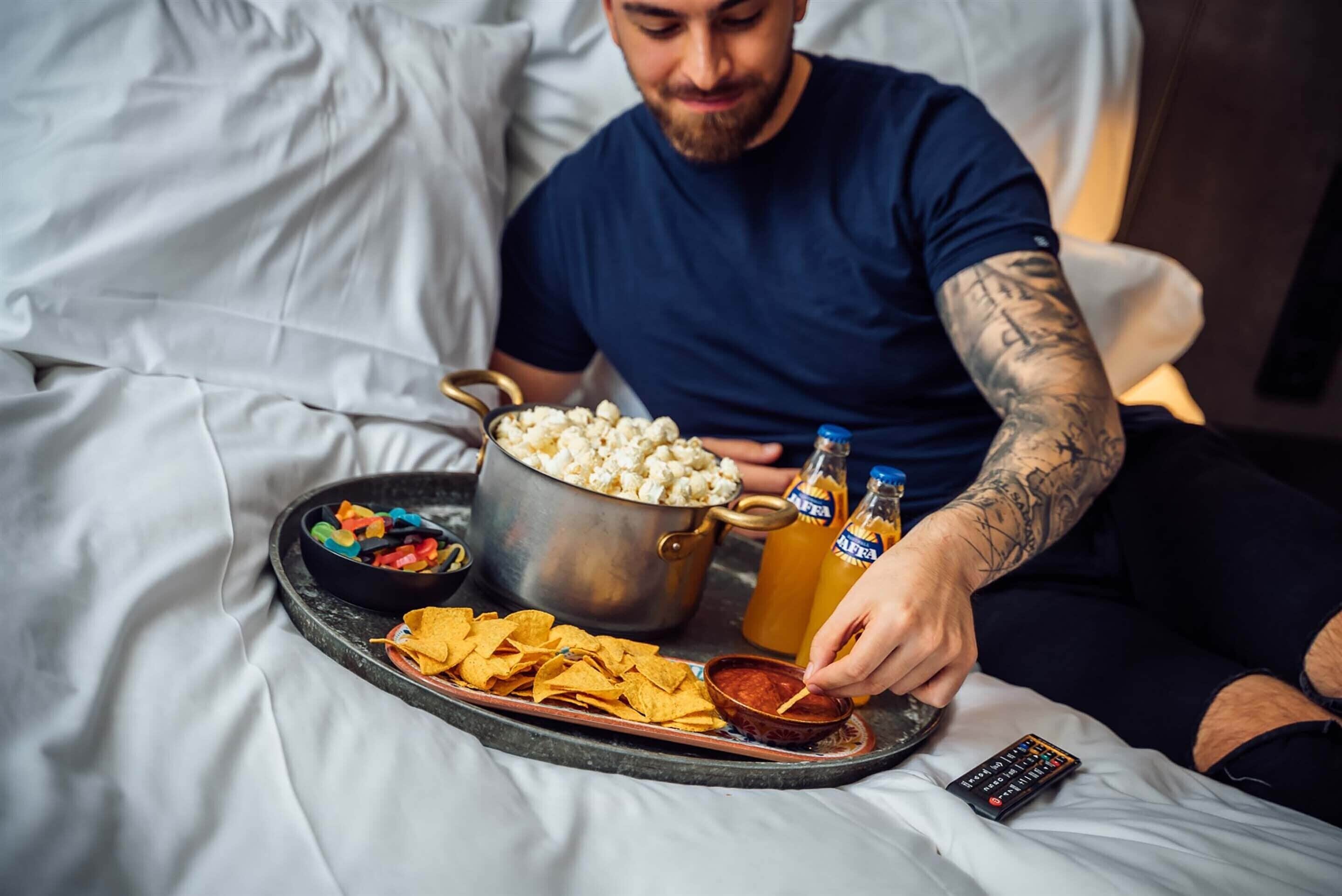 A person sitting on a bed with a tray containing popcorn, nachos, and a bowl of dip. There is a pot of popcorn and two bottles of beer on the bed.